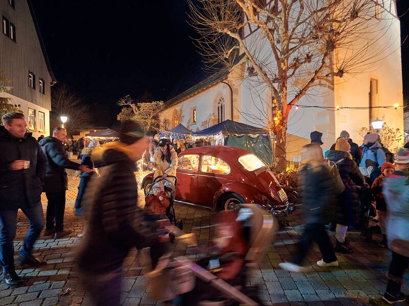 Eine festliche Straßenszene bei Nacht mit einem roten Oldtimer, umgeben von Menschen, die an einem Weihnachtsmarkt vorbeigehen.