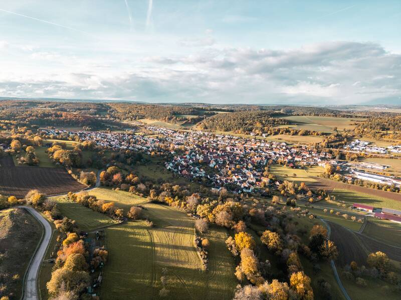 Das Bild zeigt eine Luftaufnahme einer ländlichen Kleinstadt, umgeben von Wäldern und Feldern in herbstlichen Farben.