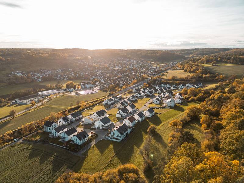 Eine ruhige Wohnsiedlung mit modernen Häusern ist von grünen Feldern und herbstlich gefärbten Bäumen in einer hügeligen Landschaft umgeben.