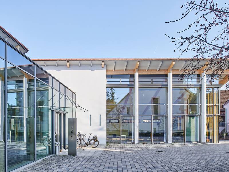 Das Bild zeigt ein modernes Gebäude mit großen Glasfronten und einem weißen Dach, umgeben von einem gepflasterten Platz und teilweise sichtbaren Bäumen.