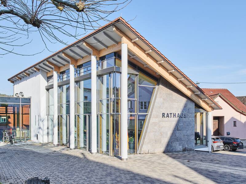 Ein modernes Rathausgebäude mit Glasfassade und spitzem Dach steht unter klarem Himmel, neben einem Baum.