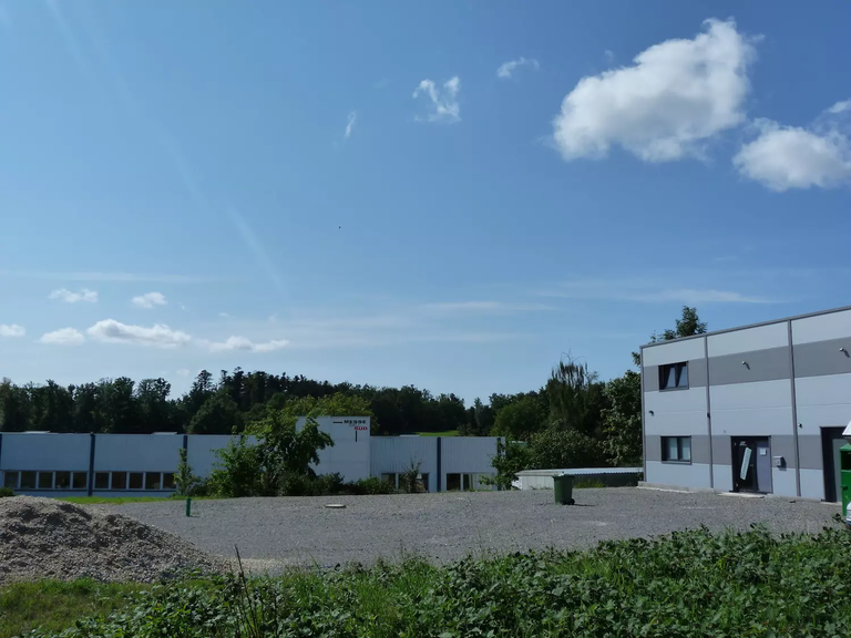Ein Industriegebäude mit weiß-grauer Fassade steht neben einem Kiesplatz unter einem blauen Himmel mit einigen Wolken.