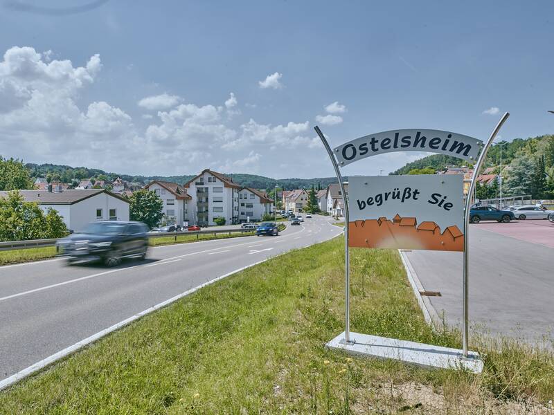 Eine Straße führt durch eine ländliche Gegend mit Häusern im Hintergrund und einem Schild, das "Ostelsheim begrüßt Sie" zeigt.