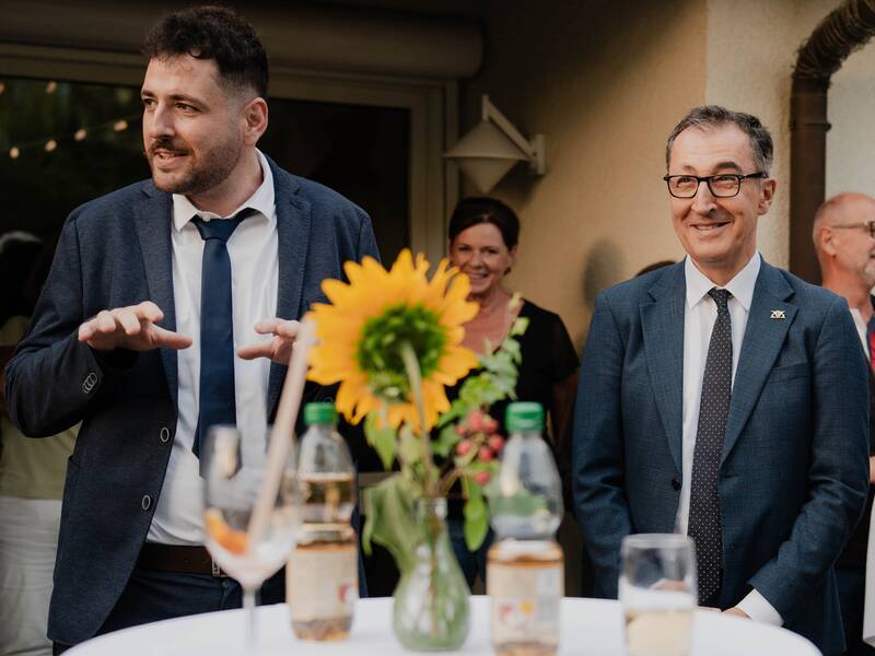 Zwei Männer in Anzügen stehen an einem Tisch mit Sonnenblumen und Getränken, umgeben von mehreren Menschen im Hintergrund.