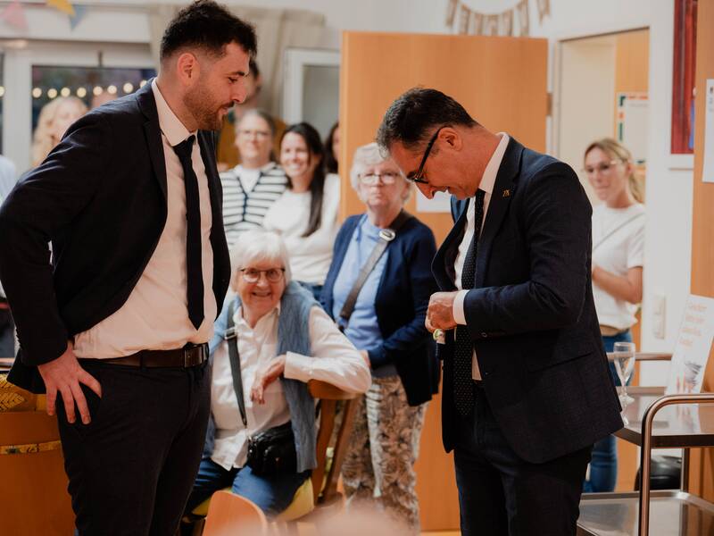 Zwei Männer in Anzügen stehen sich gegenüber in einem Raum voller Menschen, die interessiert zuschauen.