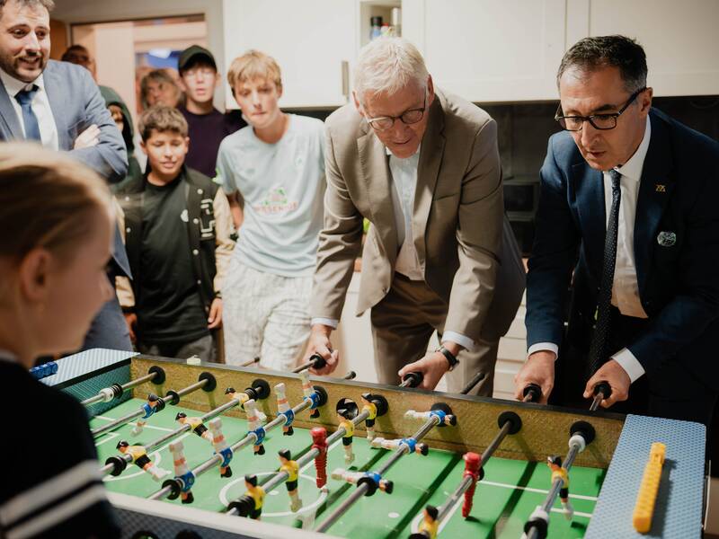 Mehrere Menschen, darunter zwei Männer in Anzügen, spielen Tischfußball, während eine Gruppe von Zuschauern interessiert zuschaut.
