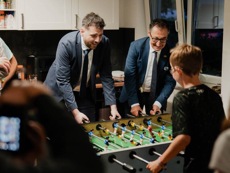 Zwei erwachsene Männer in Anzügen spielen Kicker mit einem Kind in einem Raum, während mehrere Personen zuschauen.