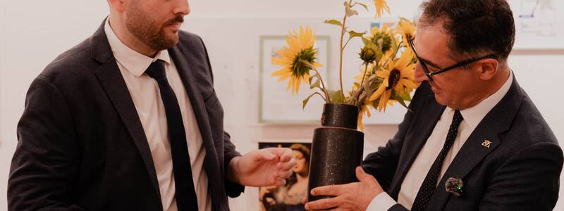 Zwei Männer im Anzug betrachten gemeinsam eine Vase mit Sonnenblumen.