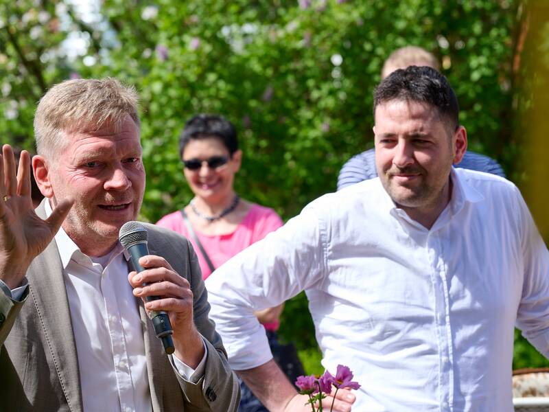 Ein Mann hält ein Mikrofon und spricht, während ein anderer Mann neben ihm lächelnd steht; im Hintergrund sind zwei weitere Personen und grüne Bäume zu sehen.