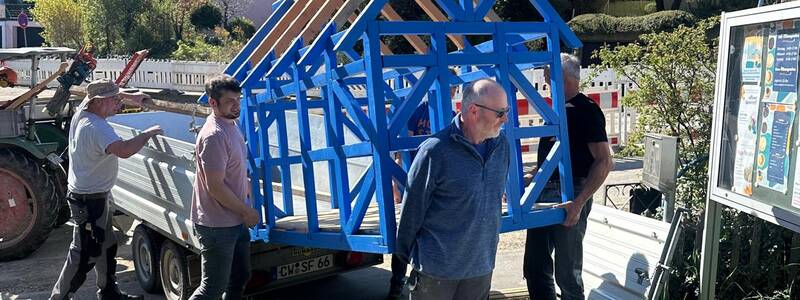 Mehrere Personen heben ein blaues Holzkonstrukt von einem Anhänger auf einen gepflasterten Gehweg in einer Wohngegend.