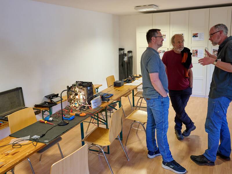 Drei Männer unterhalten sich in einem Raum mit Holztischen, auf denen Werkzeuge und ein offener Computer stehen.