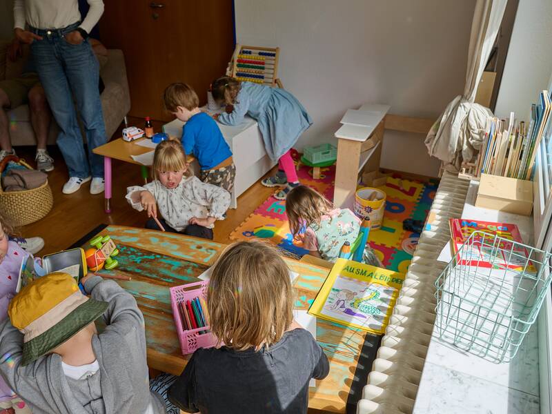 In einem sonnendurchfluteten Raum spielen mehrere Kinder mit Spielzeugen und Büchern, während Erwachsene im Hintergrund stehen.
