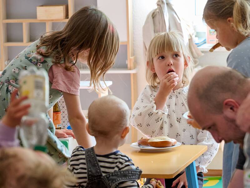 In einem hellen Raum sind mehrere Kinder und Erwachsene versammelt, wobei ein Kind an einem kleinen Tisch sitzt und isst, während die anderen um es herumstehen oder sitzen.