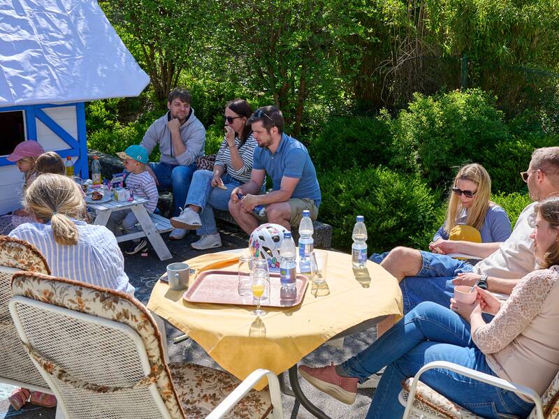 Eine Gruppe von Menschen, darunter Erwachsene und Kinder, sitzt bei sonnigem Wetter im Freien um einen Tisch, während im Hintergrund ein kleines Spielhaus steht.