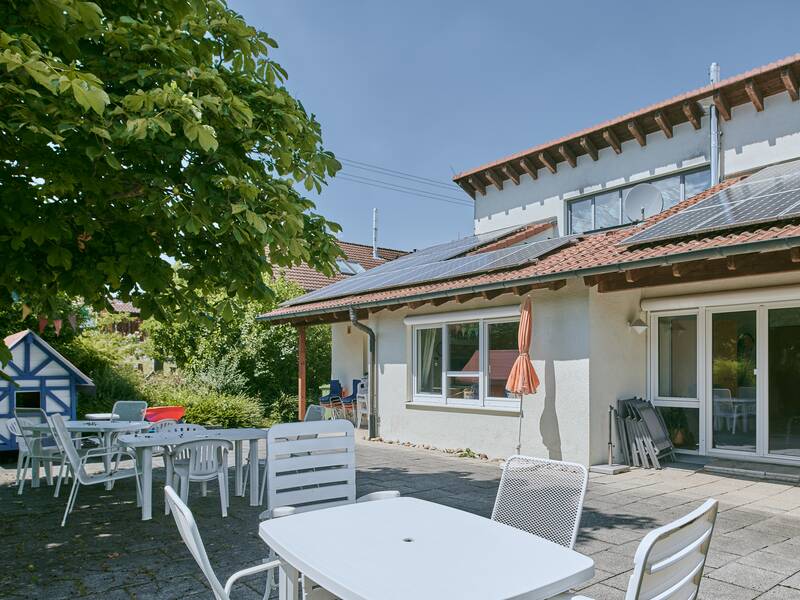 Die Aufnahme zeigt eine Terrasse mit weißen Gartenmöbeln, einem großen Baum und einem Gebäude im Hintergrund, das Solarpaneele auf dem Dach hat.