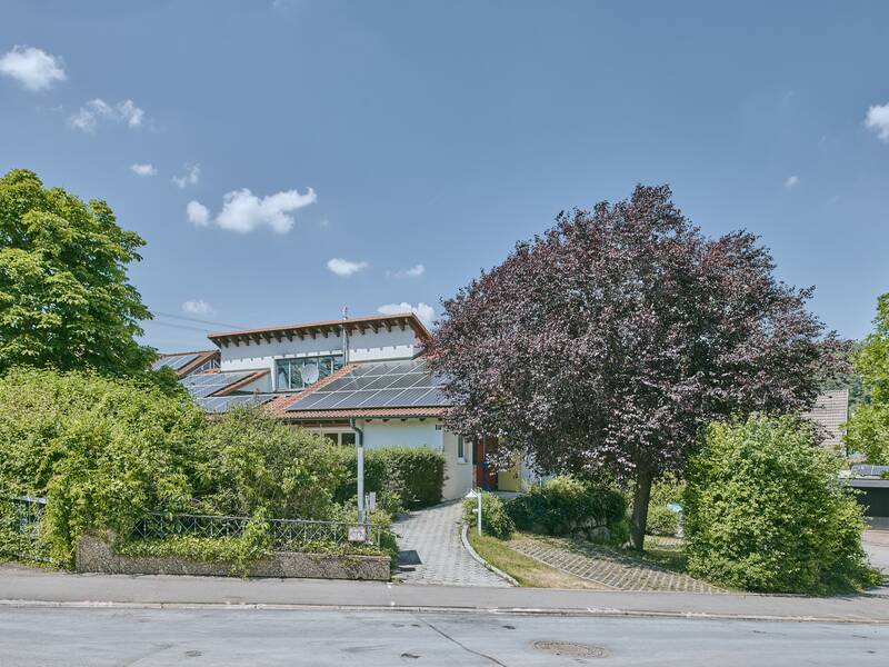 Ein modernes Haus mit Solaranlage auf dem Dach, umgeben von viel Grün und großen Bäumen, steht unter einem klaren blauen Himmel.