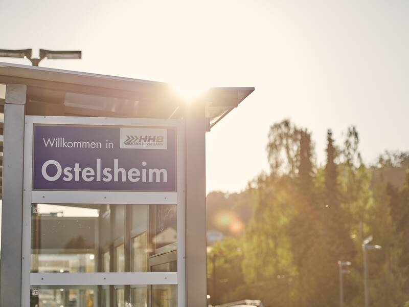 Eine Willkommensschild mit der Aufschrift "Willkommen in Ostelsheim" ist an einer Haltestelle bei Sonnenschein zu sehen.