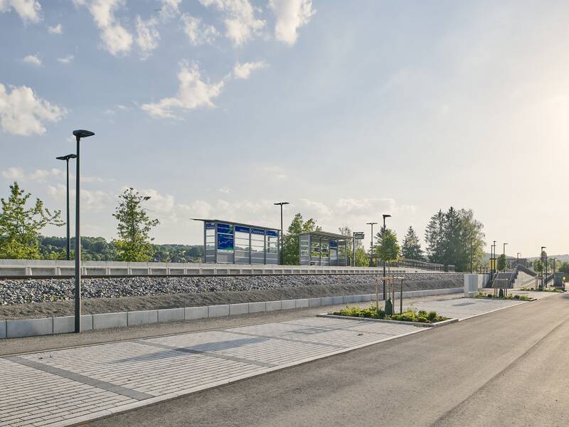 Ein moderner, leerer Bahnhof mit Bahnsteig, Gleisen und Informationsschildern, umgeben von Bäumen und einer klaren Himmelssicht.