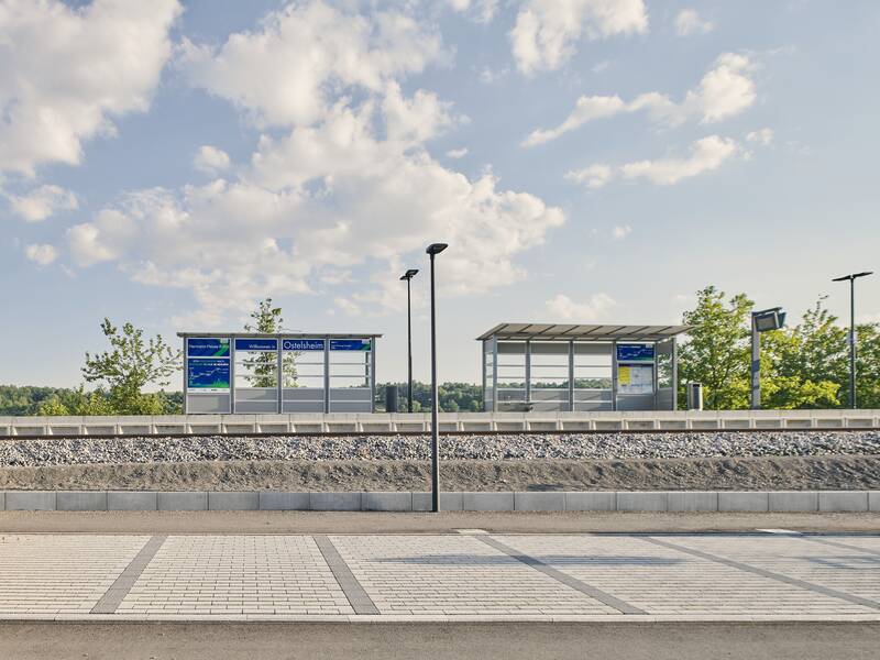 Ein leerer, moderner Bahnsteig mit einem Wartehäuschen und Informationsschildern, umgeben von Bäumen unter einem leicht bewölkten Himmel.