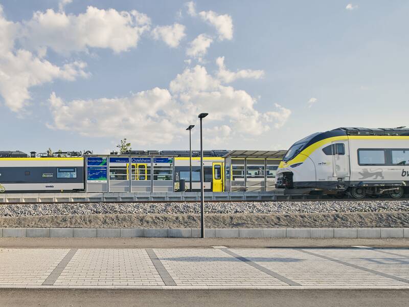 Ein moderner, gelb-grauer Triebzug steht an einem Bahnsteig mit dem Schild "Ostelsheim" unter einem blauen Himmel mit wenigen Wolken.