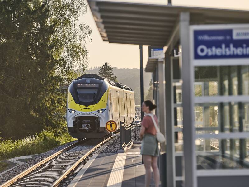 Ein gelb-weißer Zug fährt Richtung Bahnhof Ostelsheim, während eine Person am Bahnsteig wartet.