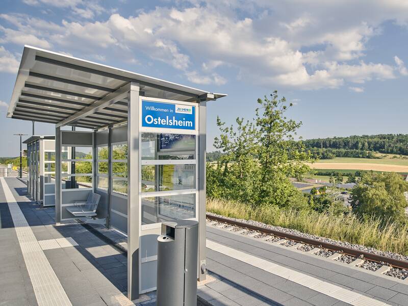 Ein moderner Bahnhof mit überdachten Wartebereichen und einem Schild, das "Willkommen in Ostelsheim" zeigt, umgeben von einer ländlichen Landschaft.