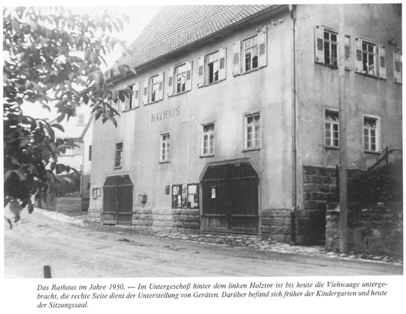 Das Bild zeigt ein altes Gebäude mit der Aufschrift "Rathaus" und mehreren Fenstern und Türen, umgeben von Bäumen.