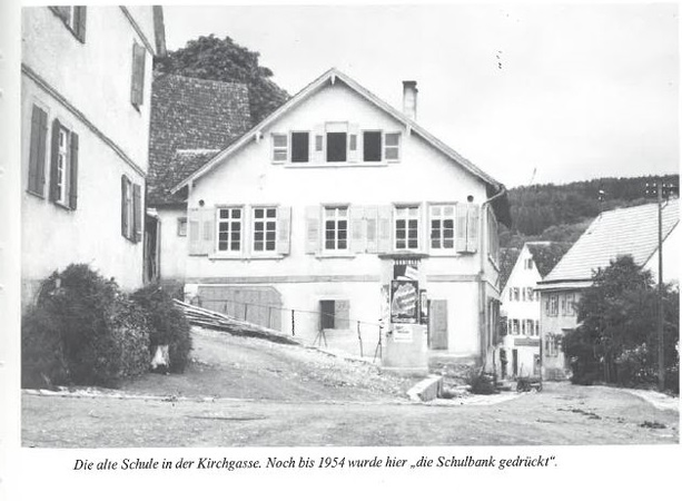 Ein altes zweigeschossiges Gebäude mit Schindeldach und Fensterläden steht am Ende einer abschüssigen Straße, umgeben von weiteren Häusern.