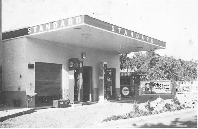 Das Bild zeigt eine historische Standard-Tankstelle mit Werbeschildern und Zapfsäulen aus einer vergangenen Zeit.