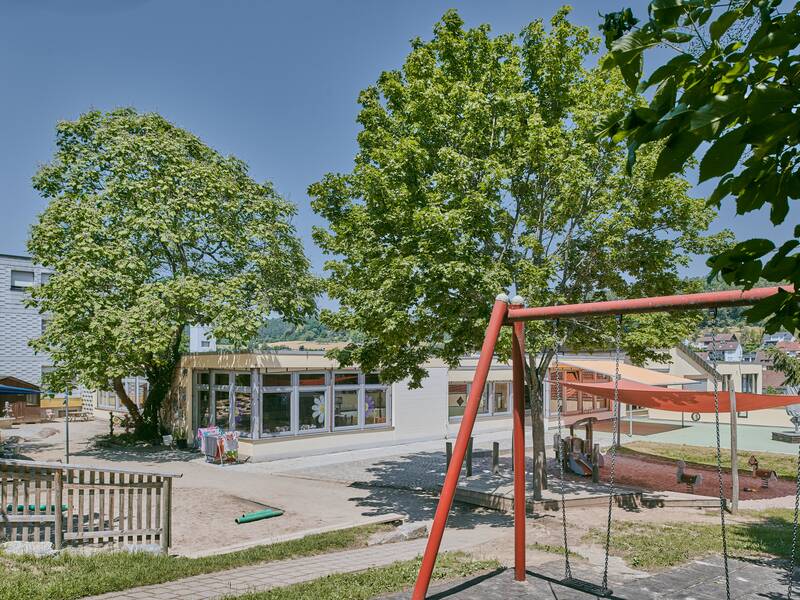 Ein Spielplatz mit Schaukeln und Bäumen ist vor einem Gebäude mit großen Fenstern zu sehen.