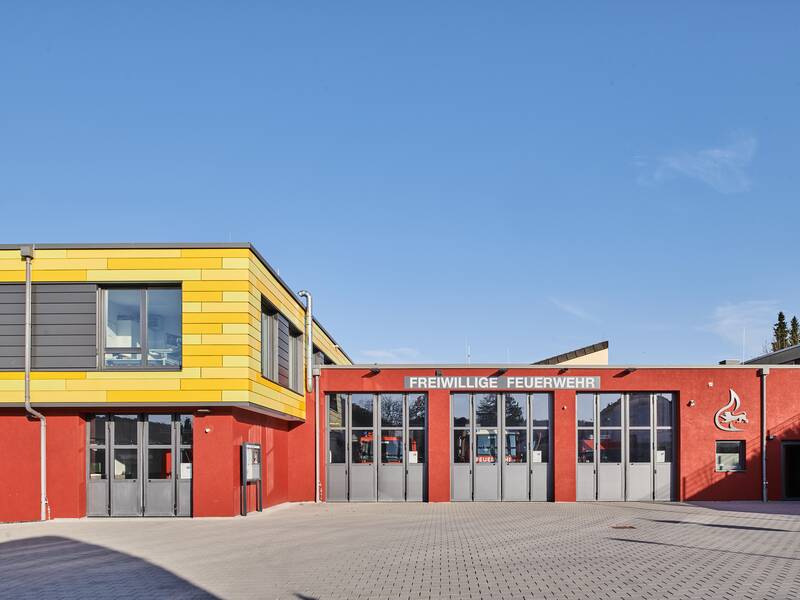 Ein modernes Gebäude der freiwilligen Feuerwehr mit einer rot-gelben Fassade und großen Fenstern bei klarem Himmel.