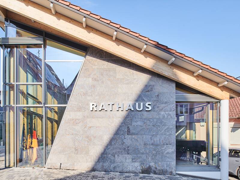 Das Bild zeigt ein modernes Rathausgebäude mit einer Glasfront und einem schrägen Dach in einem klaren blauen Himmel.