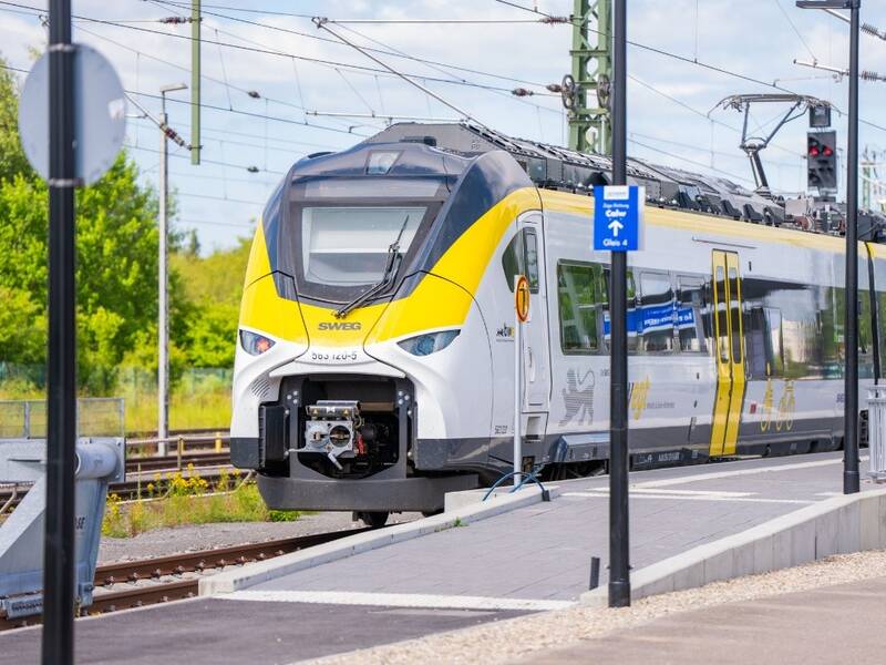 Ein moderner, gelb-weißer Regionalzug steht auf einem Bahnhofsgleis, umgeben von Oberleitungen und Vegetation im Hintergrund.
