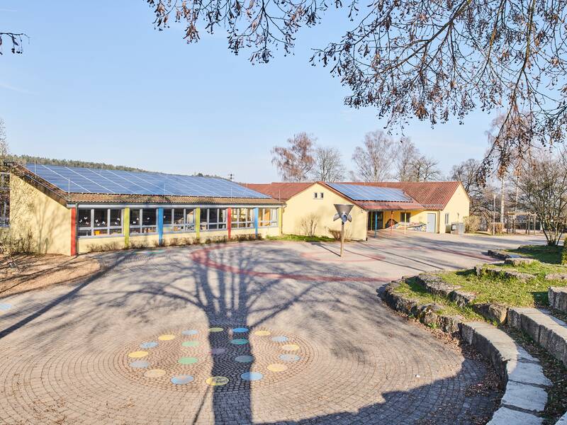 Ein Gebäude mit Solarpaneelen auf dem Dach steht vor einer gepflasterten Fläche, umgeben von Bäumen und grünem Gras.