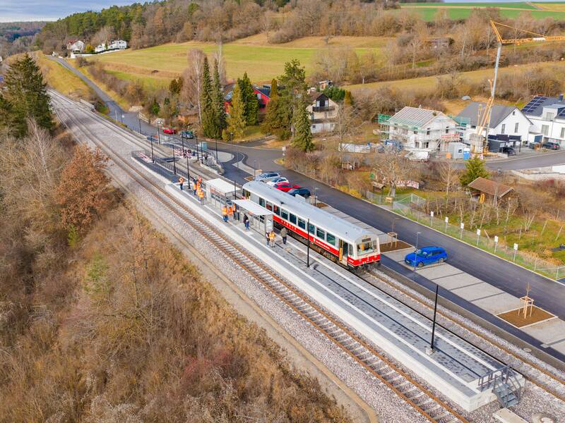 Eine Drohnenaufnahme zeigt einen Zug, der an einem modernen, ländlichen Bahnhof hält, umgeben von grünen Feldern, Bäumen und einigen Häusern.