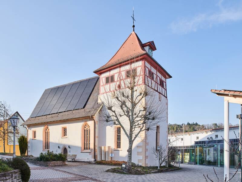 Das Bild zeigt eine historische Kirche mit einem rot-weiß gestrichenen Fachwerkturm und einem daneben stehenden Glasgebäude, umgeben von einem gepflasterten Vorplatz und einem blauen Himmel.